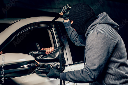 The robber in Balaclava threatening with a knife takes away a bag from the driver of a car. The concept of crime.