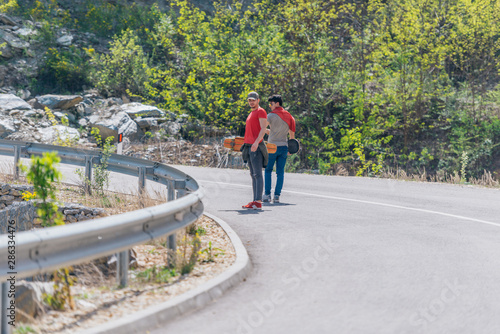 Two male longboarders carrying their longboards in their hands while climbing uphill and preparing for a downhill slide. Wearing red t-shirts  green hat  and super cool sunglasses.