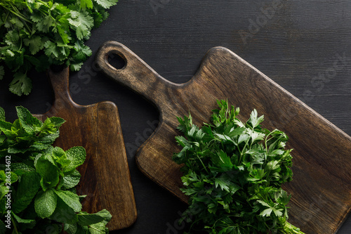 Top view of brown wooden cutting boards with parsley, peppermint and cilantro bundles on dark surface