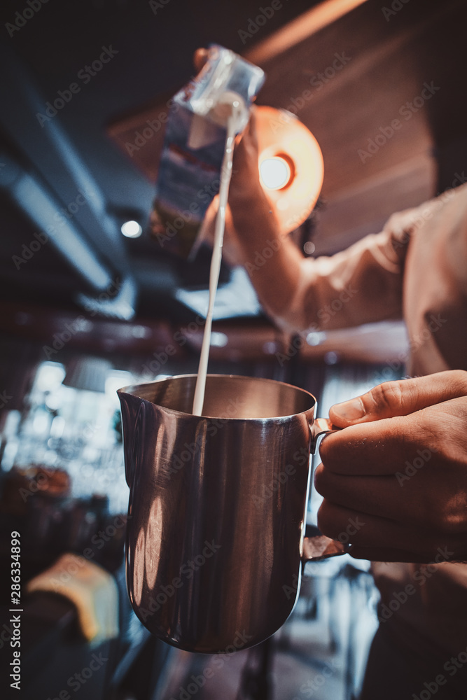 Talanted barista is pouring milk to the jug for latte or cappuchino at his cafe.