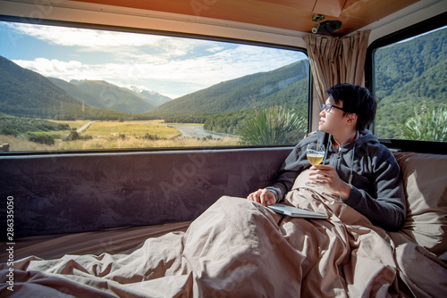 Young Asian man drinking beer after working with laptop computer on the bed in camper van looking at mountain scenery through the window. Digital nomad concept