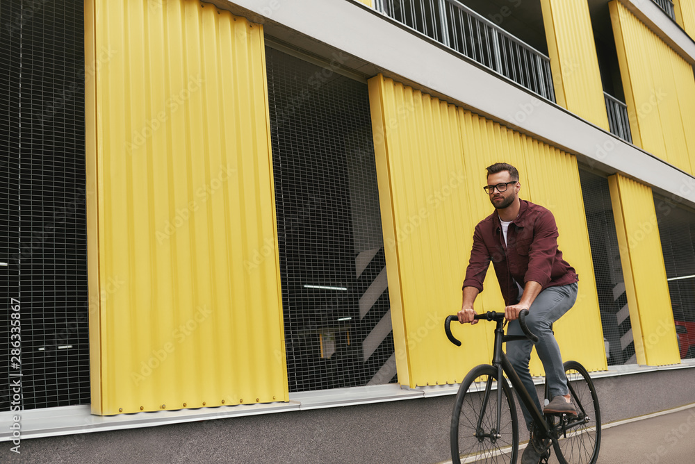 Free time. Handsome man with stubble in casual clothes riding on his bicycle along a road against yellow building