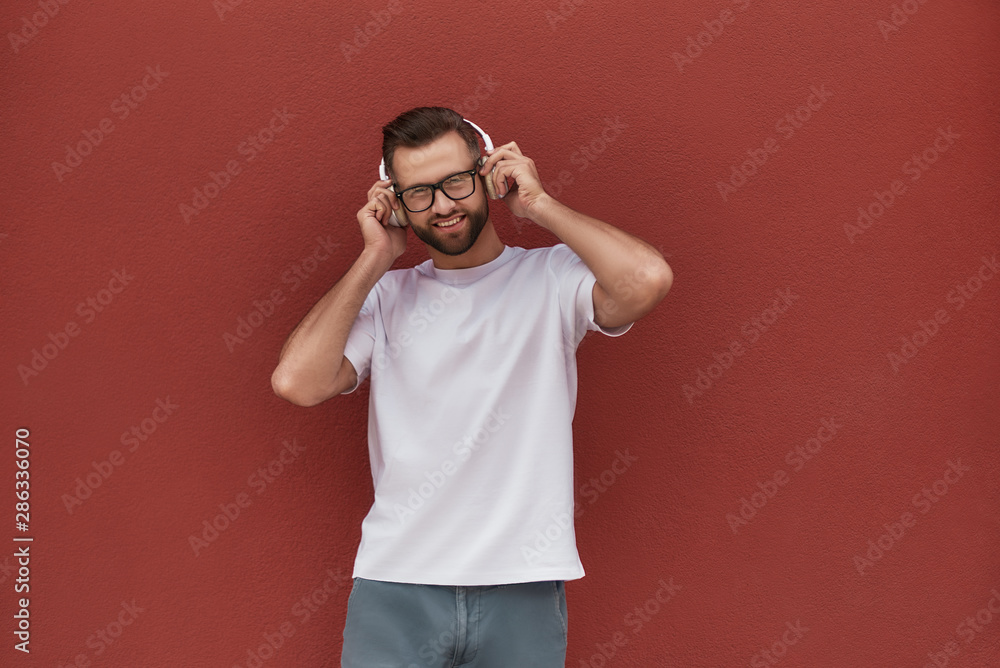 Enjoying my favourite song. Portrait of handsome and cheerful man in eyeglasses wearing headphones and listening to the music while standing against red wall outdoors Enjoying my favourite song. Portrait of handsome and cheerful man in eyeglasses wearing headphones and listening to the music while standing against red wall outdoors