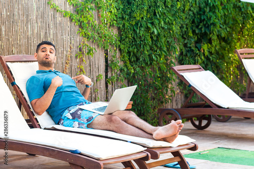 Portrait of smug handsome bearded young adult man in blue t-shirt and shorts lying on cozy sunbed with laptop on poolside and pointing finger to himself, looking at camera. Lifestyle concept, outdoor © khosrork