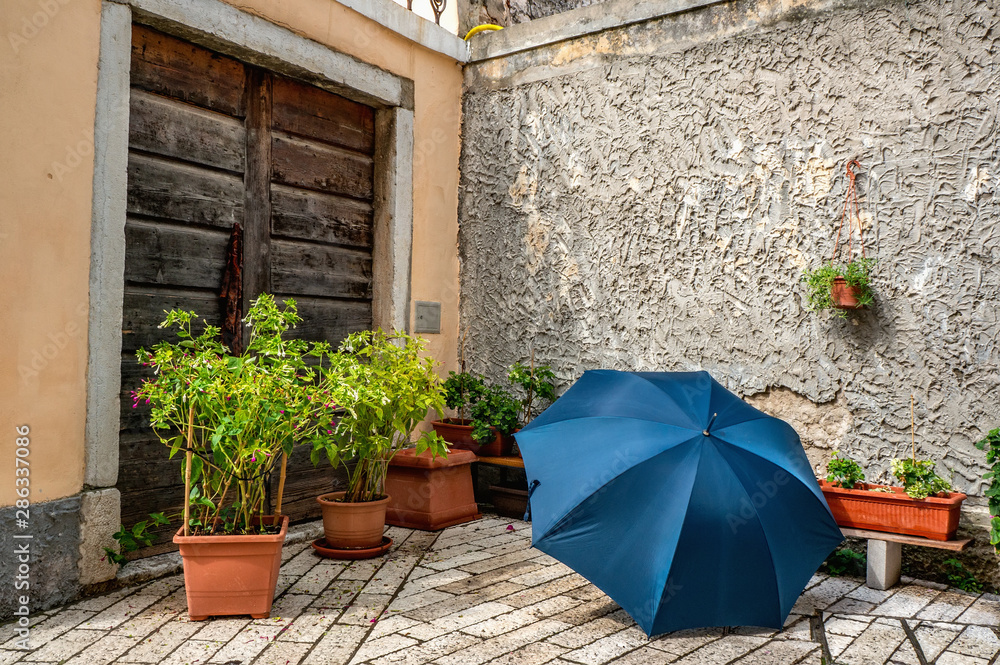 Beautiful blue umbrella lies on the cobblestones among the flowers on the background of stone walls. Design and landscaping of streets