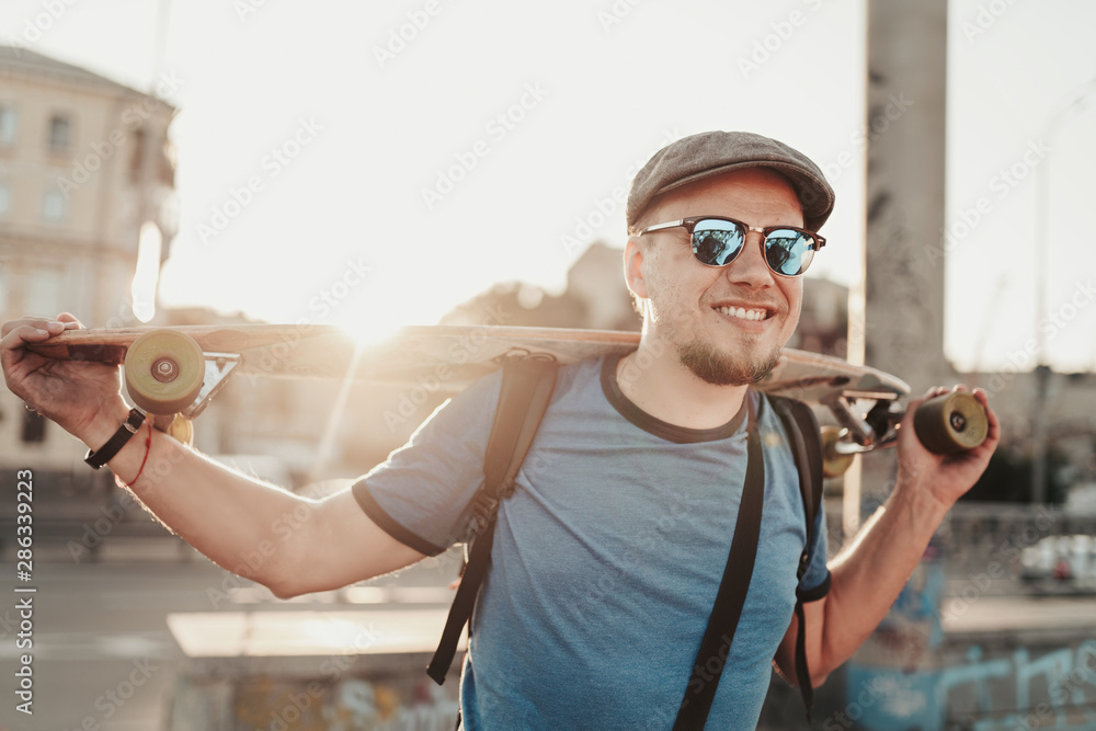 Fototapeta premium Happy hipster guy posing with skateboard in sunset in summer evening. Portrait of handsome stylish man model in blue shirt, sunglasses and hat on city street background