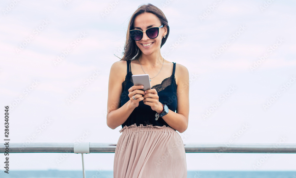 Vacation, travel, relaxation, freedom and happy time. Beautiful smiling young brunette woman in sunglasses is using her smart phone on the sea background