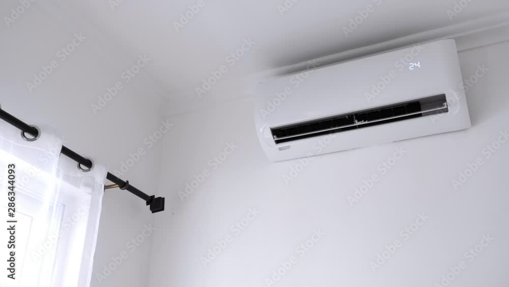 Flat air conditioner on white wall in room interior. Man pushing button on remote. Setting temperature