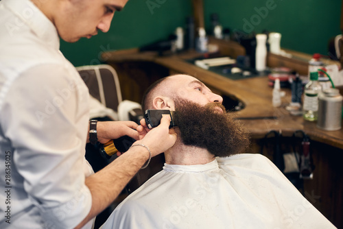 Adult client man in hairdresser chair throwing back his head during barber styling client's beard with shaver machine. Professional tools, accessories and care products on blurred background.