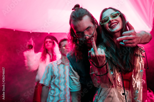 happy girl and man in sunglasses showing middle fingers in nightclub during rave party