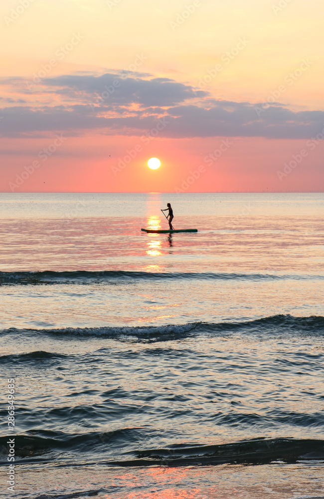 Fototapeta premium Woman's silhouette on a paddle board (SUP) at a beautiful pink pastel gradient sunset.