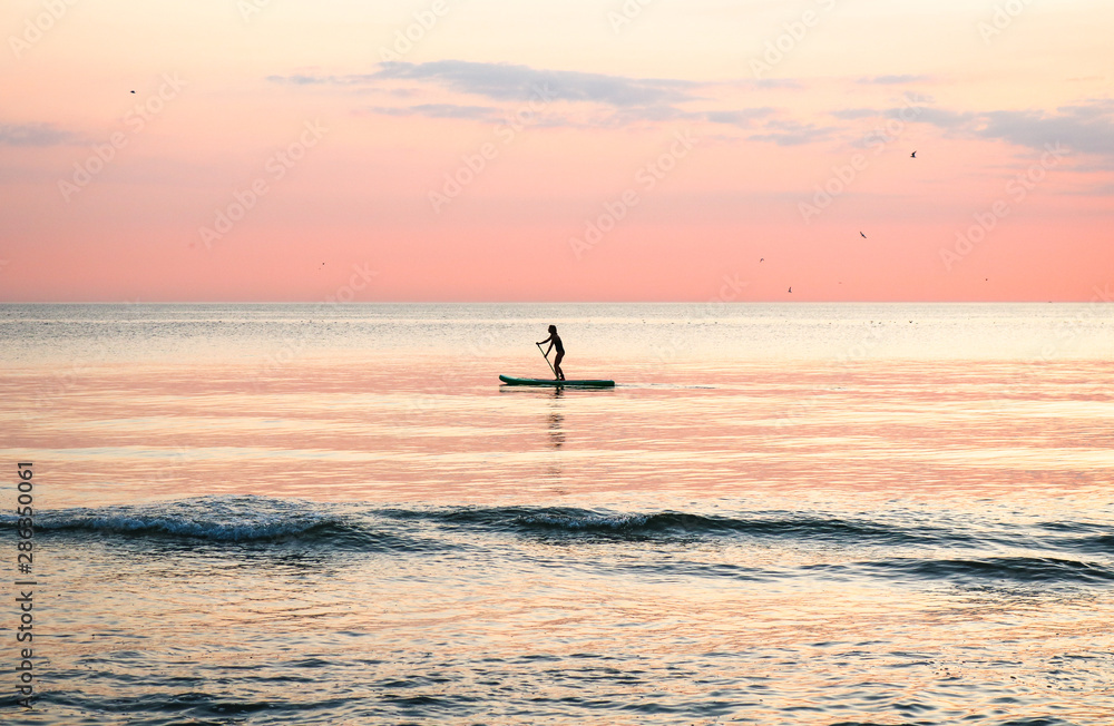 Fototapeta premium Woman's silhouette on a paddle board (SUP) at a beautiful pink pastel gradient sunset.