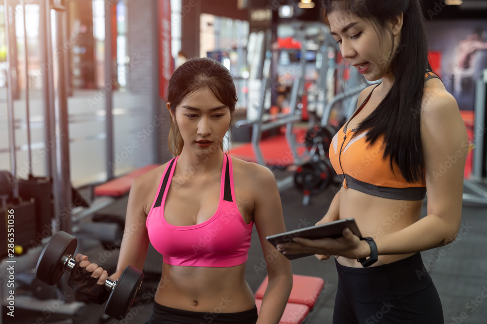Asian woman lifting a dumbbell with assistance of her personal trainer at public gym.