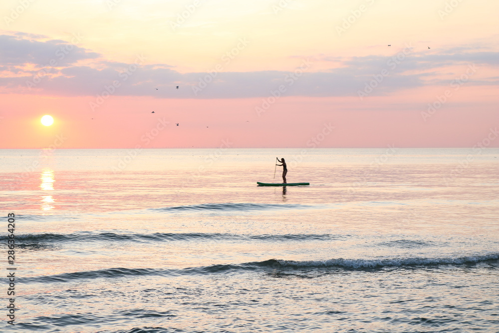 Woman's silhouette on a paddle board (SUP) at a beautiful pink pastel gradient sunset.