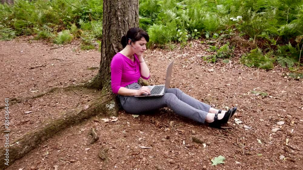 Businesswoman talking on her phone whilst working on a laptop in the woods Businesswoman talking on her phone whilst working on a laptop in the woods