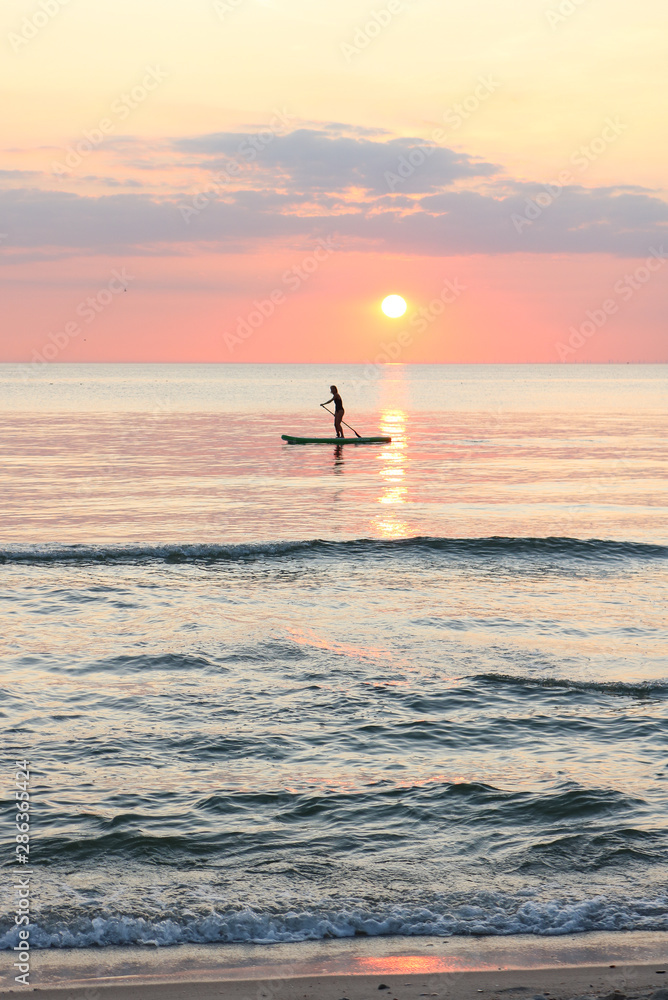 Fototapeta premium Woman's silhouette on a paddle board (SUP) at a beautiful pink pastel gradient sunset.