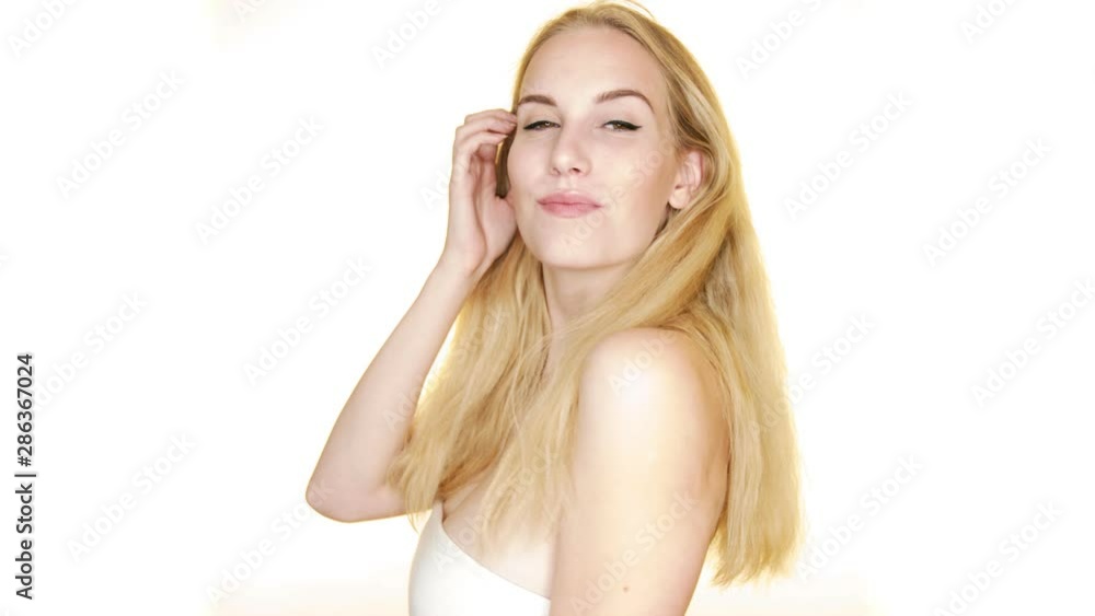 Against a plain white studio background, a young woman with blonde hair looks over her shoulder and smiles
