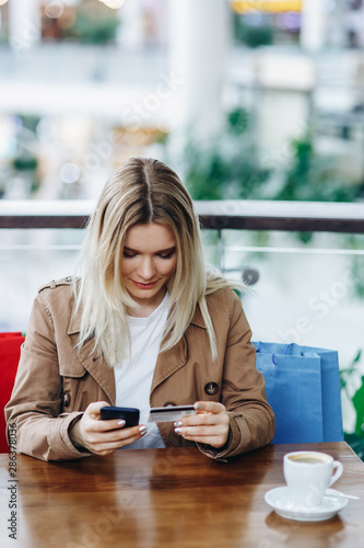 The girl spent a lot of money. Blonde woman in brown cloak with shopping bags in cafe at shopping mall. Lady buying online with a credit card and smartphone. Copy space above the head