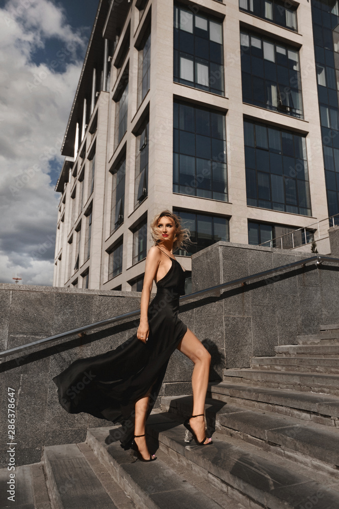 Beautiful, sexy and fashionable blonde girl with perfect body in black evening dress posing at urban background Beautiful, sexy and fashionable blonde girl with perfect body in black evening dress posing at urban background