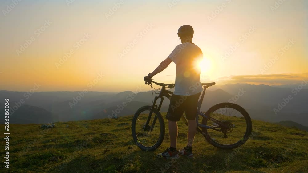 SLOW MOTION, SUN FLARE: Cross country biker stands on top of a hill and watches the sunset. Cool mountain bike dude rests and relaxes as he observes the picturesque golden lit countryside in Slovenia. SLOW MOTION, SUN FLARE: Cross country biker stands on top of a hill and watches the sunset. Cool mountain bike dude rests and relaxes as he observes the picturesque golden lit countryside in Slovenia.