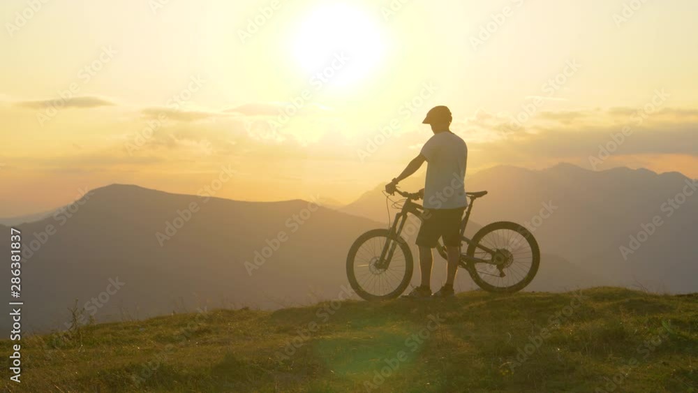 SLOW MOTION DRONE LENS FLARE COPY SPACE: Male tourist observes the stunning golden landscape before mountain biking in the morning. Cross country cyclist watches the sunset after riding in mountains. SLOW MOTION DRONE LENS FLARE COPY SPACE: Male tourist observes the stunning golden landscape before mountain biking in the morning. Cross country cyclist watches the sunset after riding in mountains.