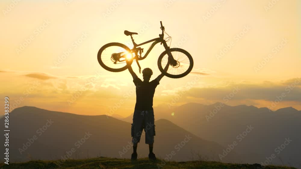 SILHOUETTE, SUN FLARE, SLOW MOTION, COPY SPACE: Unrecognizable fit male tourist holds bike above head after biking up a grassy hill at sunrise. Bicycle rider celebrates an awesome cross country trip.