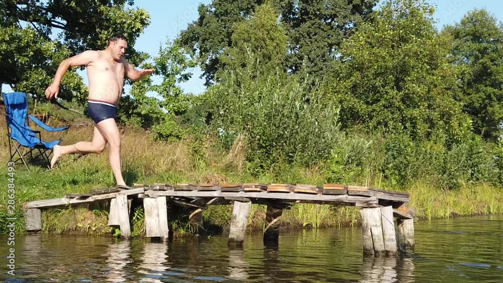 A man jumps into the lake from a wooden pavement. A man dives into the lake from a wooden bridge. A man jumps into the lake from a wooden pavement. A man dives into the lake from a wooden bridge.