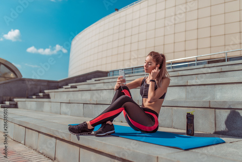 Beautiful woman sits on mat, fitness in summer city, outdoors, for yoga after workout, relaxation, active lifestyle, shaker energy drink protein, headphones online application smartphone, free space.