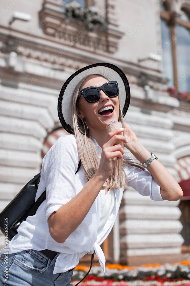 Beautiful young woman in hat and glasses walking on the streets of a European city. Lovely girl traveling in Europe. Perfect blonde European woman eating blueberry icecream cones and enjoy life..