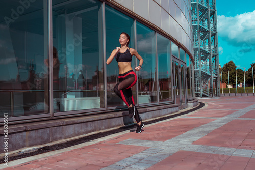 beautiful and young woman, in summer morning in city runs in jump, background is glass windows of building, active lifestyle, fitness in nature, free space, sportswear leggings top, tanned figure.