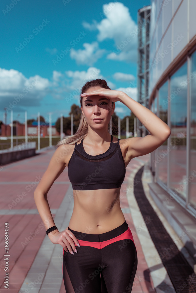 A sports woman jogging in morning, looks into distance, covers her hand with an eye from bright sun, sportswear with black leggings top, in summer in city, active lifestyle fitness jogging