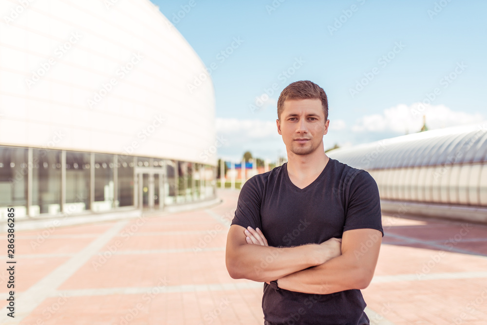 Male athlete, stands in the summer in the city against the background of the building, sports an active lifestyle, posing, black T-shirt, muscular arms. Motivation and confidence
