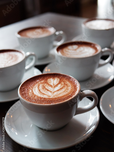 Set of freshly brewed cappuccino cups with milk foam latte art served on bartend table in cozy city cafe lit with natural window light
