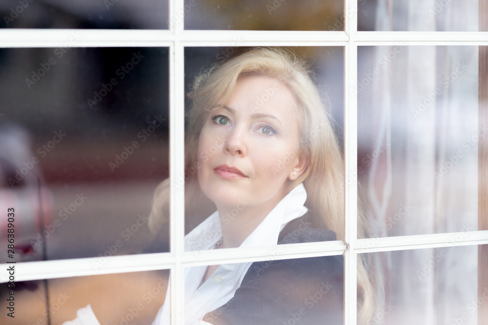 A horizontal portrait of Caucasian woman with long blond hair behind a window glass, dreamy 