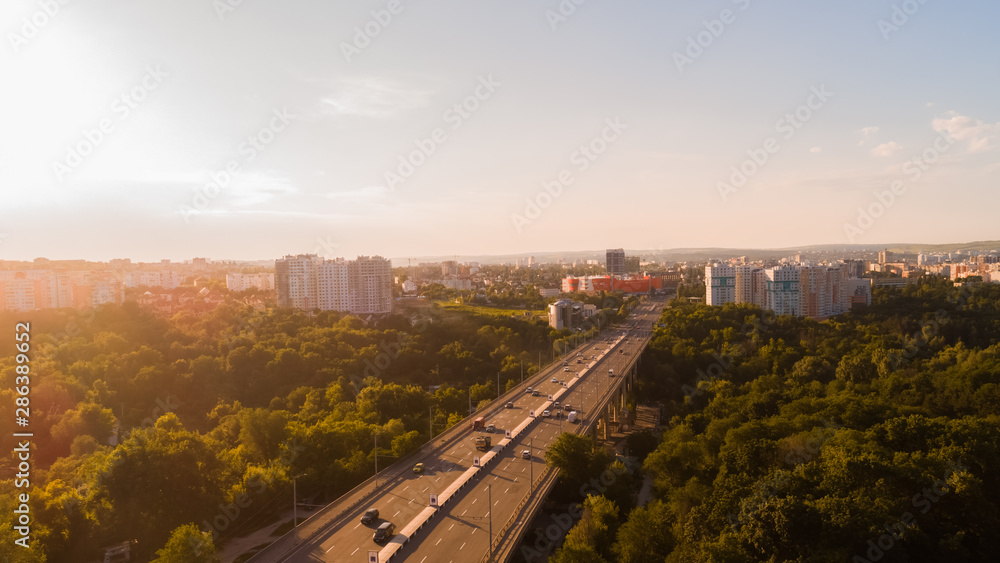 Aerial view of a highway road located in the middle of a green forest leading towards a city, during a majestic sunrise.