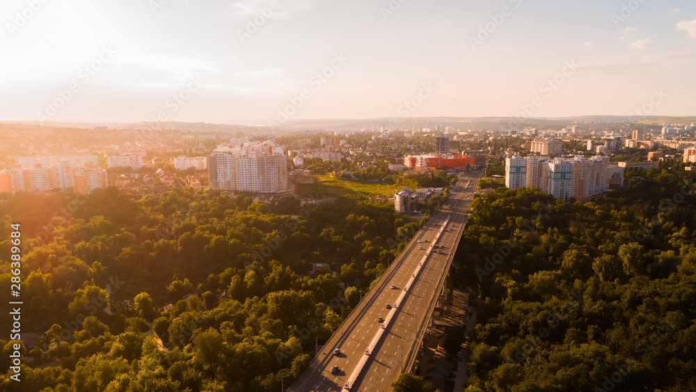 Aerial view of a highway road located in the middle of a forest leading towards a big city, during a majestic sunrise.