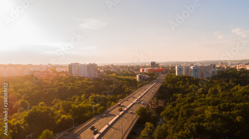 Aerial view of a highway road located in the middle of a green forest leading towards a city, during a majestic sunrise.