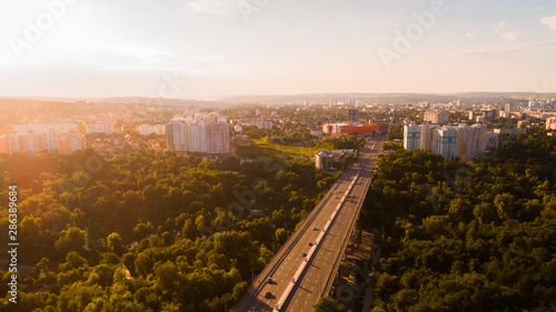 Fototapeta Naklejka Na Ścianę i Meble -  Aerial view of a highway road located in the middle of a forest leading towards a big city, during a majestic sunrise.