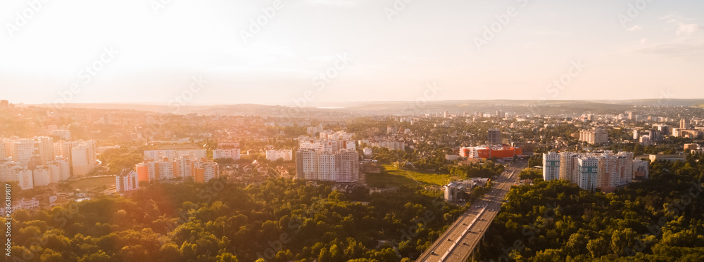 Aerial view of a highway road located in the middle of a green forest leading towards a city, during sunrise.