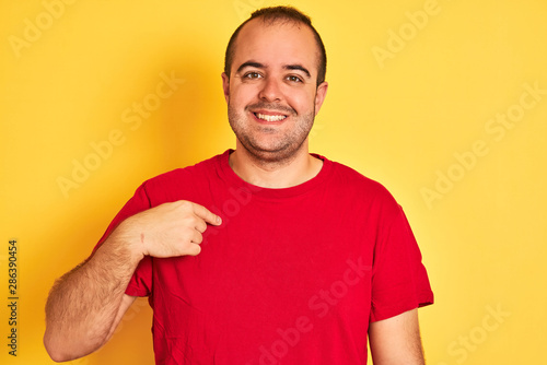 Young man wearing red casual t-shirt standing over isolated yellow background with surprise face pointing finger to himself