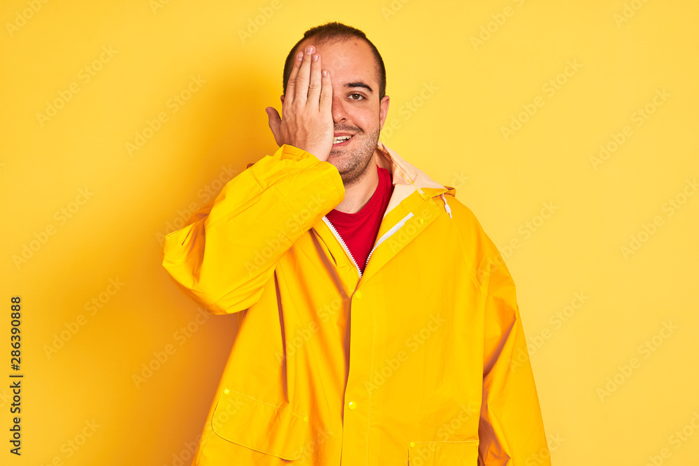 Young man wearing rain coat standing over isolated yellow background covering one eye with hand, confident smile on face and surprise emotion. Young man wearing rain coat standing over isolated yellow background covering one eye with hand, confident smile on face and surprise emotion.