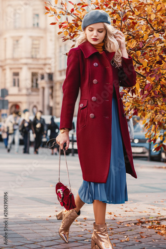 Street style autumn fashion concept: full-length portrait of elegant lady wearing burgundy color woolen coat, light blue pleated skirt, beret, metallic color ankle boots, holding small quilted bag