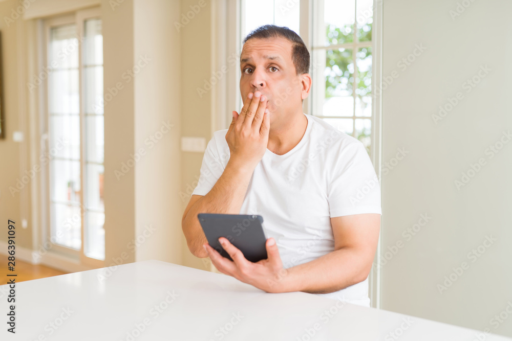 Middle age man using digital table at home cover mouth with hand shocked with shame for mistake, expression of fear, scared in silence, secret concept