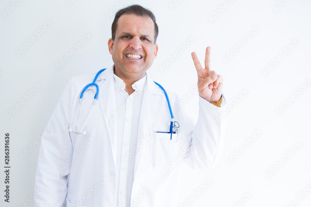 Middle age doctor man wearing stethoscope and medical coat over white background smiling with happy face winking at the camera doing victory sign. Number two. Middle age doctor man wearing stethoscope and medical coat over white background smiling with happy face winking at the camera doing victory sign. Number two.