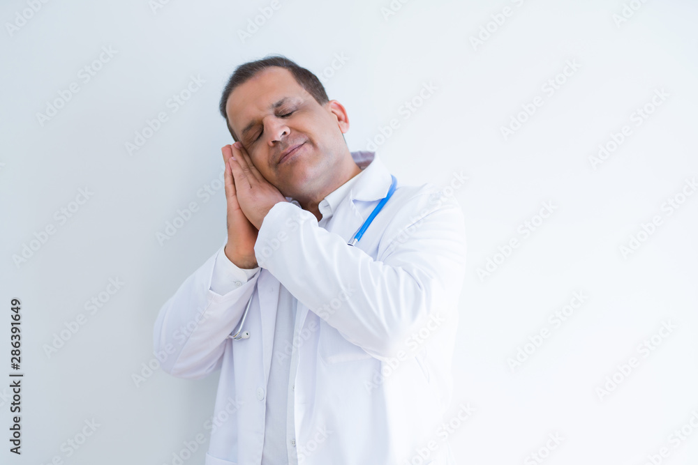 Middle age doctor man wearing stethoscope and medical coat over white background sleeping tired dreaming and posing with hands together while smiling with closed eyes. Middle age doctor man wearing stethoscope and medical coat over white background sleeping tired dreaming and posing with hands together while smiling with closed eyes.