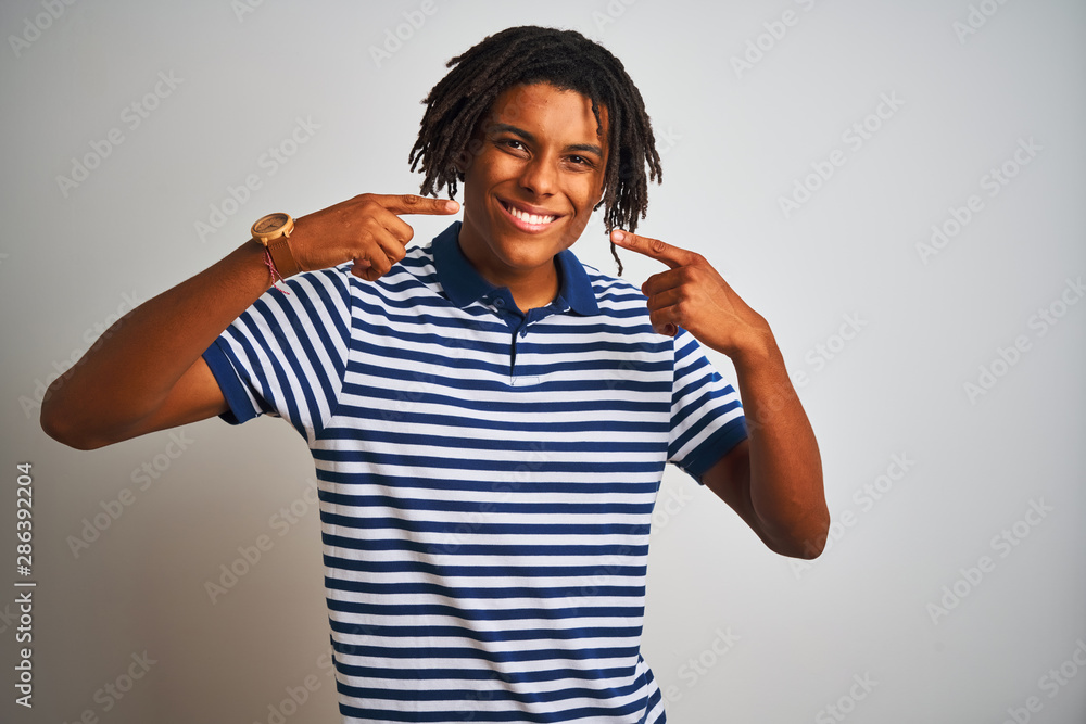 Afro man with dreadlocks wearing striped blue polo standing over isolated white background smiling cheerful showing and pointing with fingers teeth and mouth. Dental health concept. Afro man with dreadlocks wearing striped blue polo standing over isolated white background smiling cheerful showing and pointing with fingers teeth and mouth. Dental health concept.