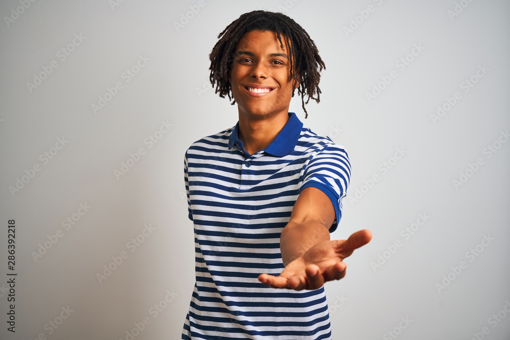 Obraz premium Afro man with dreadlocks wearing striped blue polo standing over isolated white background smiling cheerful offering palm hand giving assistance and acceptance.