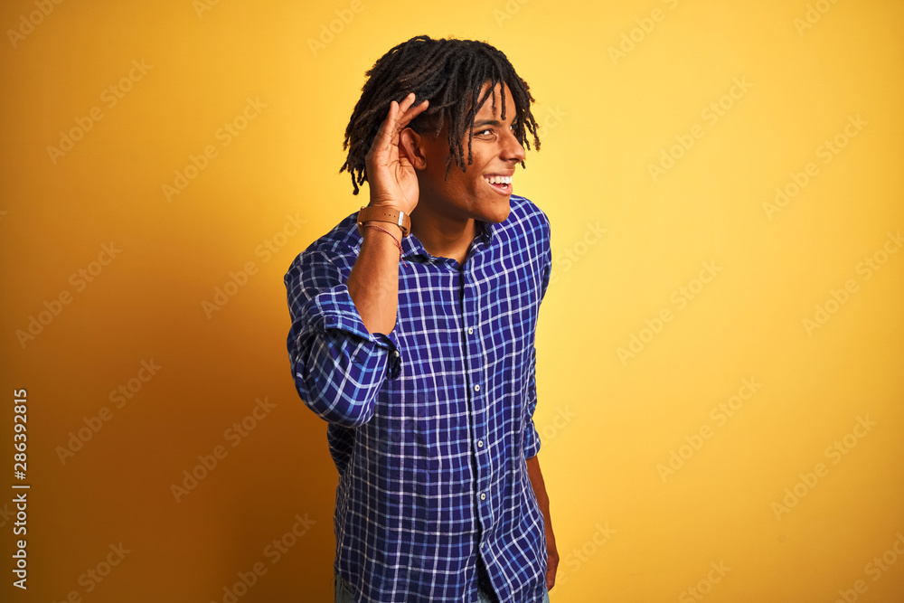 Afro man with dreadlocks wearing casual shirt standing over isolated yellow background smiling with hand over ear listening an hearing to rumor or gossip. Deafness concept. Afro man with dreadlocks wearing casual shirt standing over isolated yellow background smiling with hand over ear listening an hearing to rumor or gossip. Deafness concept.