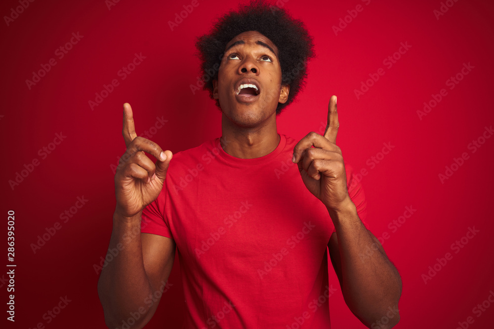 Young american man with afro hair wearing t-shirt standing over isolated red background amazed and surprised looking up and pointing with fingers and raised arms. Young american man with afro hair wearing t-shirt standing over isolated red background amazed and surprised looking up and pointing with fingers and raised arms.