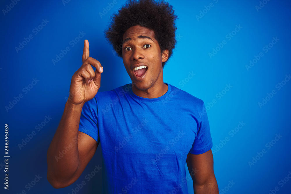 African american man with afro hair wearing t-shirt standing over isolated blue background pointing finger up with successful idea. Exited and happy. Number one. African american man with afro hair wearing t-shirt standing over isolated blue background pointing finger up with successful idea. Exited and happy. Number one.